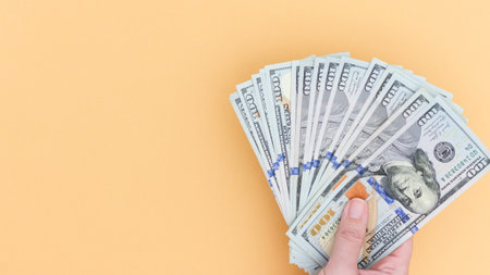 A hand holds a fan of one hundred dollar bills against a light orange background. Symbolizes wealth and finance, perfect for visuals related to investment and economic prosperity.の写真素材