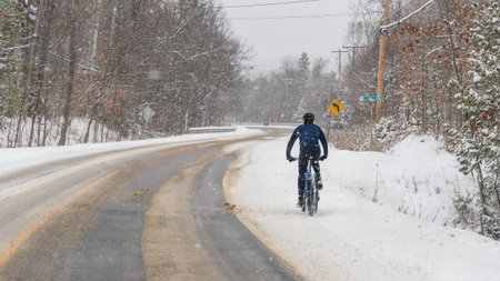 Gatineau QC, Canada - November 30, 2025: Cyclist in black jacket rides a blue bike on snowy road, trees and street sign visible in winter weatherのeditorial素材