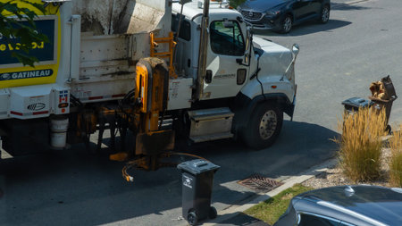 Gatineau QC, Canada - September 10, 2025: Garbage truck is collecting waste on residential street surrounded by trees and parked vehicles, emphasizing urban sanitation effortsのeditorial素材