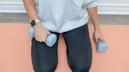 Woman wearing a gray sweatshirt kneels on an exercise mat, holding light blue dumbbells in each...