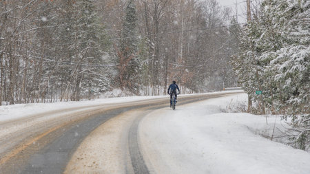 Gatineau QC, Canada - November 30, 2025: Bicyclist in blue jacket rides on snowy road surrounded by trees during snowstorm, winter landscape visibleのeditorial素材