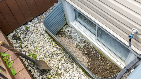 Gravel and soil mixture fills a window well, bordered by decorative stones and a shovel,...