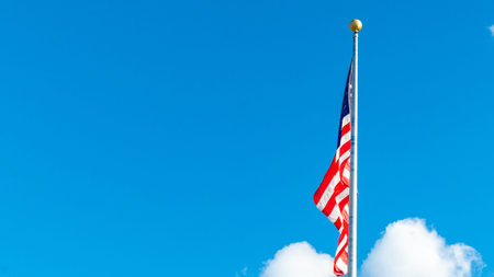 American flag is waving in the wind against a clear blue sky with soft white clouds, representing...