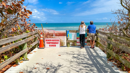 Miami FL, USA - March 01, 2026: Two individuals observe the ocean from a wooden boardwalk, with a sign stating BEACH ACCESS TEMPORARILY CLOSED and coastal vegetation surrounding the areaのeditorial素材