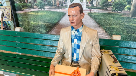 Savannah GA, USA - February 28, 2026: Forrest Gump statue depicted in a light-colored suit and blue checkered shirt, seated on a green bench, holding a gift box with an orange ribbon in a park environmentのeditorial素材