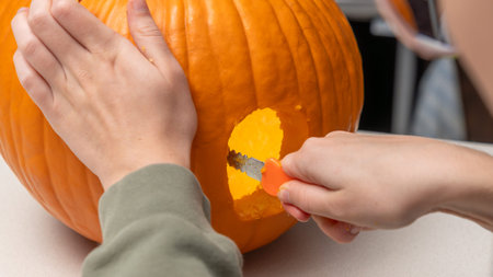 Individual using a small knife to carve a pumpkin for Halloween, revealing the bright orange flesh and hollowed interior, with hands positioned carefully on the pumpkin's surface