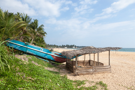 Colorful Sri Lankan fishing boat on sandy beach with palm trees in the background and a little hut in the foreground.の写真素材
