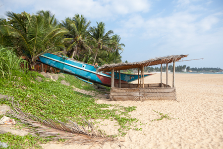 Colorful Sri Lankan fishing boat on sandy beach with palm trees in the background and a little hut in the foreground.の写真素材