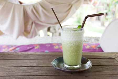 Fresh organic green tea drink with ice cubes served in a glass with a brown drinking straw and a handmade plate underneath. Hammock and natural surroundings in the background.の写真素材