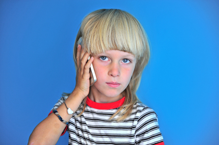 Portrait of blonde long hair boy talking by mobile phone on deep blue background at homeの写真素材