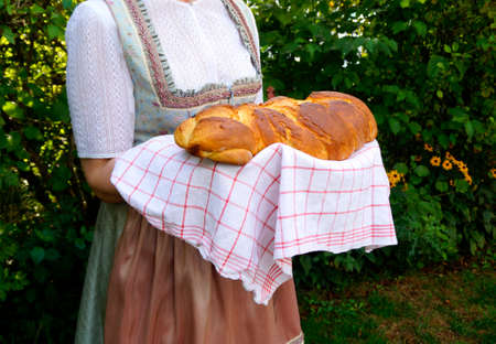 woman in beautiful traditional Bavarian or also Austrian dirndl dress (or Tracht) holding delicious freshly baked home-made loaf of rustic german bread called Zopf with brown crustの写真素材