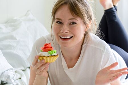 Joyful woman lying on the bed and holds cakeの写真素材