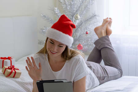 Christmas online congratulations. Joyful girl in santa hat is lying on the bed and making video call using tabletの写真素材