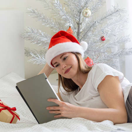 Christmas online congratulations. Smiling woman in santa hat is lying on the bed and making video call using tabletの写真素材