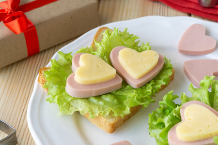 Breakfast for valentines day. Sandwich with sausage, cheese and lettuce on white plateの写真素材