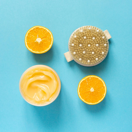 Orange anti-cellulite cream and brush for dry massage on blue background, top view.