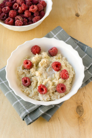 Milk barley porridge with butter and raspberries in white bowl on wooden backgroundの写真素材