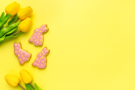 Easter flat lay. Homemade cookies in the shape of bunnies with tulips on yellow background, space...