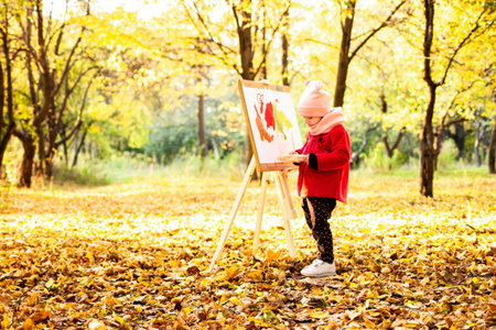 A little girl in a red coat paints a watercolor on the easel in the park against the background of the autumn landscape.の写真素材