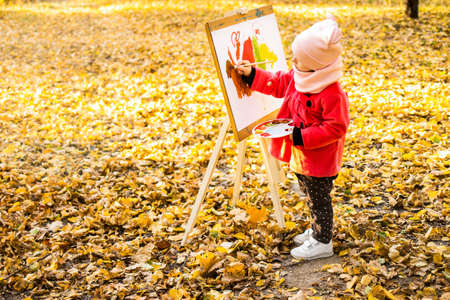 A little girl in a red coat paints a watercolor on the easel in the park against the background of the autumn landscape.の写真素材