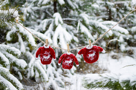 Knitted toy red sweaters with deer hanging on a rope on spruce branches in the winter forest during snowfall. The atmosphere of Christmas and New Year.の写真素材