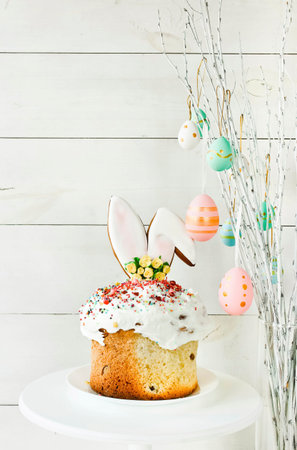 Easter cake stands on a white plate on a white wooden background. The branches of the trees...