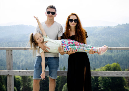 A happy young family of European appearance smiles against the background of mountains. Mom, Dad, and little daughter are happy with the trip to the mountains. The concept of travel, tourism, a happy family, and a happy childhood.の写真素材