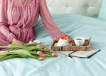 A woman is eating breakfast in bed. A tray with food on the bed. A cake and a white cup with coffee with pink tulips on a tray. Romantic breakfast in the bed. Special events.の写真素材