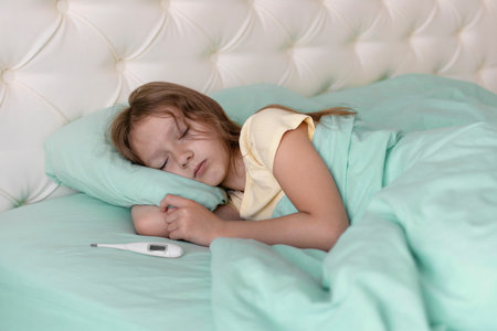 A little girl is sleeping in bed, with a thermometer next to her pillow. Concept of illness season...