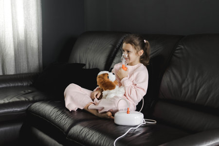 A little girl sits on a black leather sofa at home, holding a stuffed toy and using a nebulizer...