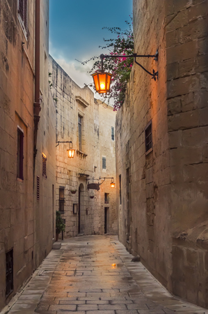 Mdina, Malta: narrow street paved with setts in medieval town with limestone walls and lantern lights in the early eveningの写真素材