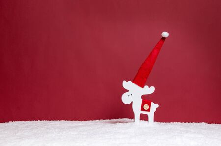 White wooden reindeer in Santa's hat standing alone in snow against red background. Minimalist Christmas concept. Merry Christmas greeting card. Copy-space for text.の写真素材