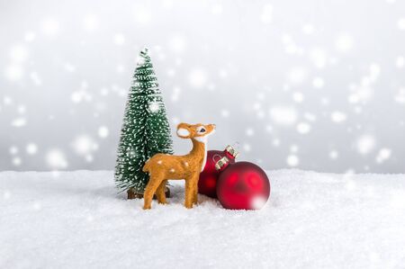 Miniature reindeer standing near fir tree and red Christmas ornaments under falling snow. Christmas holiday theme against light festive background with snowflakes and copy-spaceの写真素材