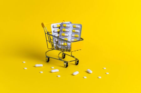 Shopping cart with blister packs and surrounded with white pills on yellow background. Creative concept of healthcare and safe online shopping for medicine at pharmacy on coronavirus quarantine.の写真素材