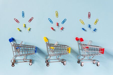 Colorful supermarket shopping trolley carts on blue background with yellow, red and blue clips and pins above. Shopping for office supplies, commerce and sale concept. Flat lay minimal compositionの写真素材