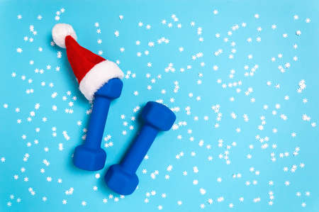 Christmas sport flat lay with dumbbells in red Santas hat on blue background. Christmas and new year holiday concept for fitness, workout and healthy lifestyle. New year resolution to exercise more.の写真素材