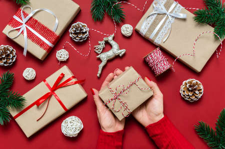 Female hands holding Christmas gift on red background. Festive holiday composition with woman hands, present boxes, pine tree, baubles and decoration. Winter holiday celebration. Top view, flat lay.の写真素材
