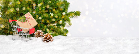 Shopping for Christmas presents. Red shopping cart with gift in snow, fir tree branches and vibrant...