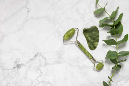 Green jade roller and gua sha stone for facial massage and eucalyptus branch on marble background. Home beauty and selfcare accessories. Face roller for anti age wrinkle treatment. Top view, flat lay.の写真素材
