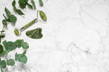 Green jade roller and gua sha stone for facial massage and eucalyptus branch on marble background. Home beauty selfcare accessories. Face roller for anti age wrinkle treatment. Top view, copy spaceの写真素材