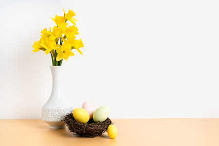 Colorful pastel easter eggs in nest and bouquet of daffodil flowers in white vase on beige table and white background. Happy Easter and holiday preparation. Festive decorationの写真素材