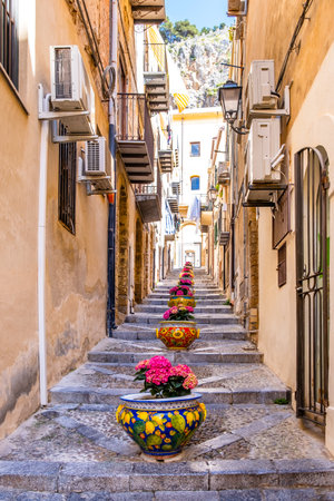 Narrow street in the old town of Cefalu, medieval village on Sicily island, Italy. Flower pots with traditional sicilian decoration. La Rocca cliff. Popular tourist attraction in Province of Palermoの写真素材