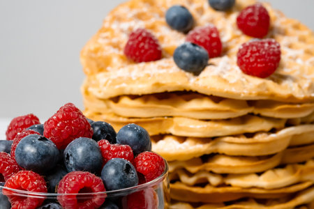 Close up of mixed raspberries and blueberries and Belgian waffle in background. Healthy delicious summer breakfast with fruit. Stack of homemade round waffles and berries. Selective focusの写真素材