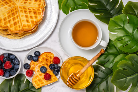Belgian waffles, berries, honey and cup of tea: delicious summer breakfast from above. Heart-shaped waffle and mix of raspberries and blueberries as topping on marble background. Flat lay, top viewの写真素材