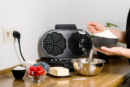 Making Belgian waffles. Female hands add flour into metal bowl to prepare dough in home kitchen. Batter ingredients: flour, eggs, butter and sugar to cook round waffles with berries in waffle makerの写真素材