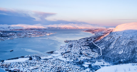 Aerial view of Tromso, Norway at sunset in winter from Storsteinen mountain. Snowy roofs of Tromso and Tromsdalen, fjord and snow capped mountains in background.の写真素材