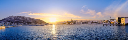 Panorama of Tromso harbor at sunset in winter, Norway. Scandinavian buildings and boats in port of Tromso and Storsteinen mountain. Snowy winter landscape in arctic circle town in far northern Norway.の写真素材