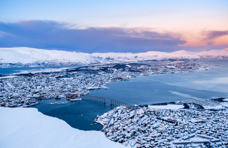 Aerial view of Tromso, Norway at sunset in winter from Storsteinen mountain. Tromsoya island with snowy roofs of Tromso downtown, harbor, Sandnessundet Bridge over Fjord and mountains in background.の写真素材