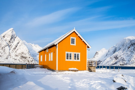 Traditional yellow rorbu house and Olstind mountain peak in Sakrisoy fishing village on Lofoten islands, Norway. Winter landscape with snowy mountains and Scandinavian cottageの写真素材