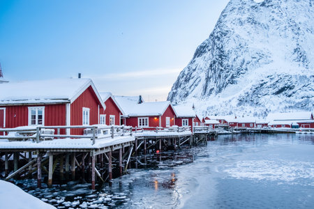 Reine village with traditional rorbu houses on Lofoten islands in winter. Scandinavian landscape with red wooden fishermen cabins on frozen sea water and snowy Reinebringen mountain in northern Norwayの写真素材
